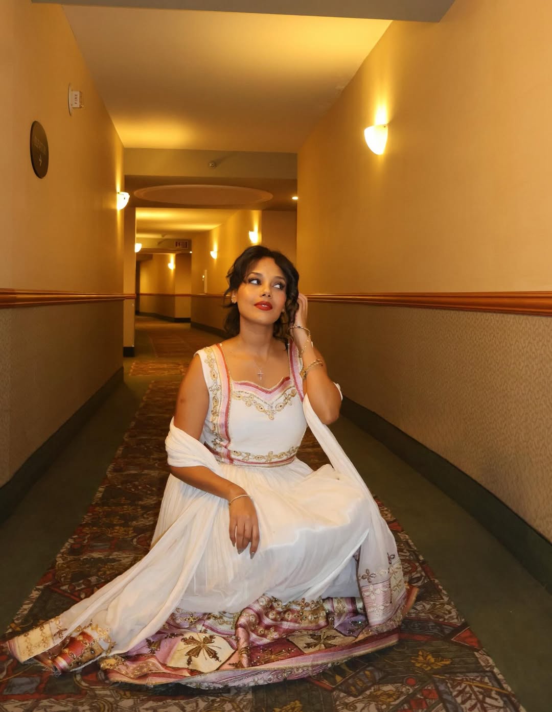 Woman in a white Ethiopian traditional dress  sitting on a patterned rug in a hallway.