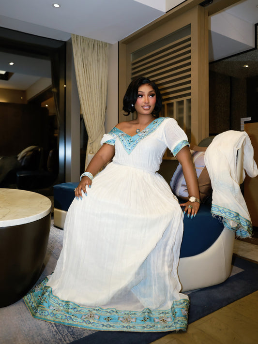 A woman wearing a white maxi  Ethiopian dress  with turquoise blue embroidery and a matching draped scarf, sitting on a modern blue and cream lounge chair in a hotel room.