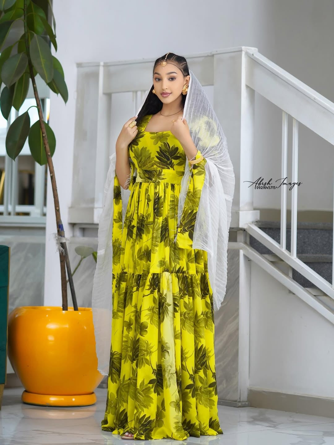Woman in a Yellow Habesha Shifion standing indoors with a plant and staircase in the background.