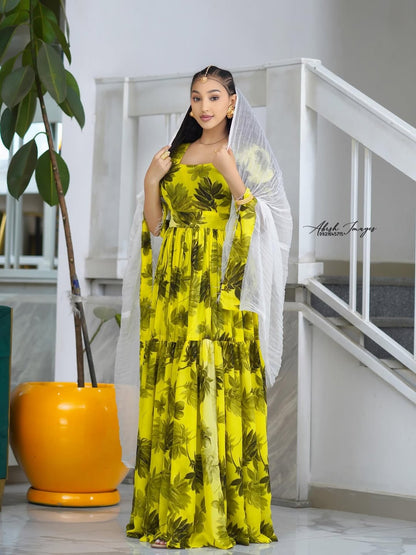 Woman in a Yellow Habesha Shifion standing indoors with a plant and staircase in the background.