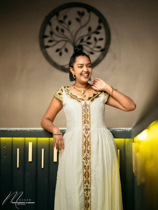 Woman in a white and gold Gold Habesha Kemis standing indoors with decorative wall and lighting in the background.