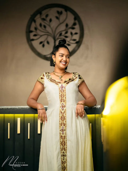 Woman in a white and gold traditional outfit standing indoors with decorative wall in the background.