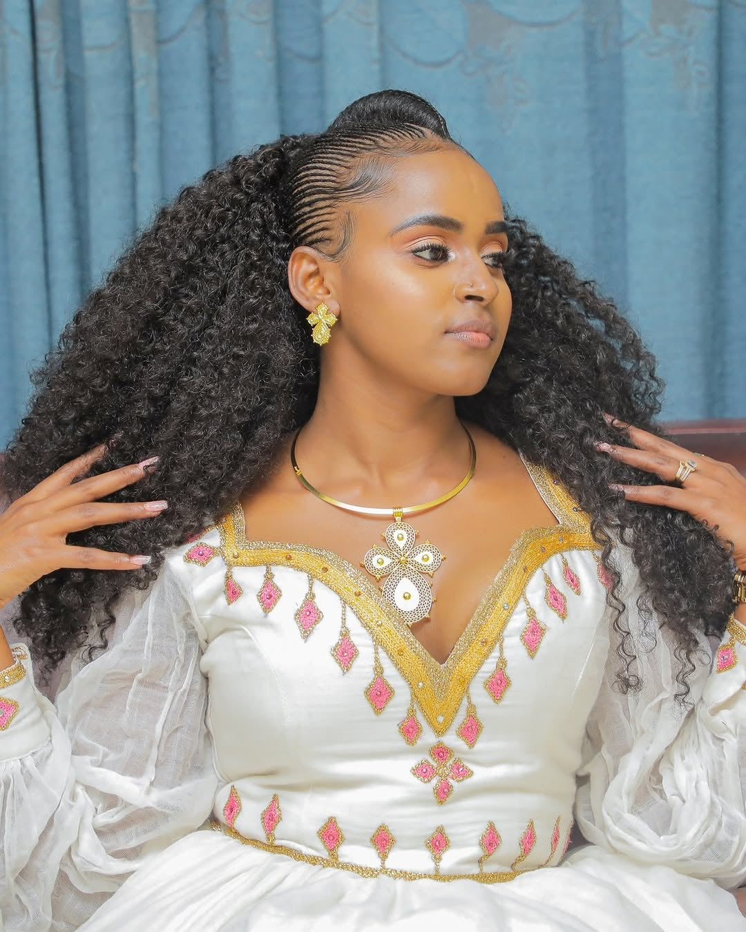 Woman with styled hair wearing a white Habesha Dress with pink and gold details against a blue curtain background.