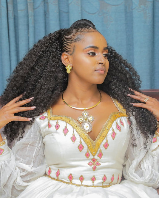 Woman with styled hair wearing a white Habesha Dress with pink and gold details against a blue curtain background.