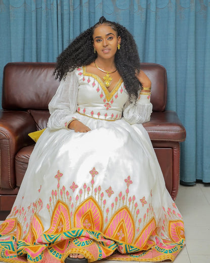 Woman in a white Habesha Dress with colorful patterns sitting on a brown couch against a blue curtain background.