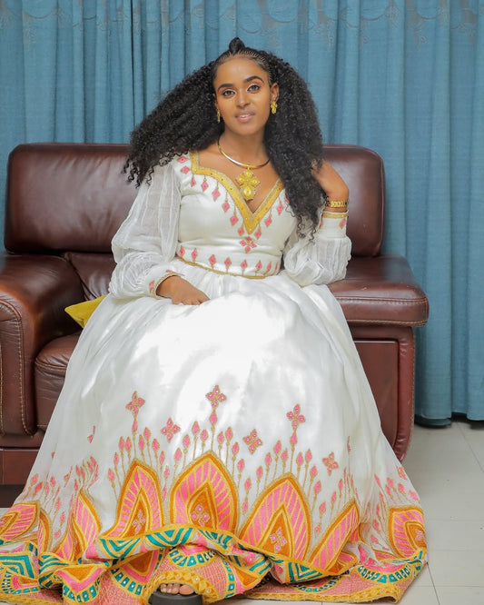 Woman in a white Habesha Dress with colorful patterns sitting on a brown couch against a blue curtain background.