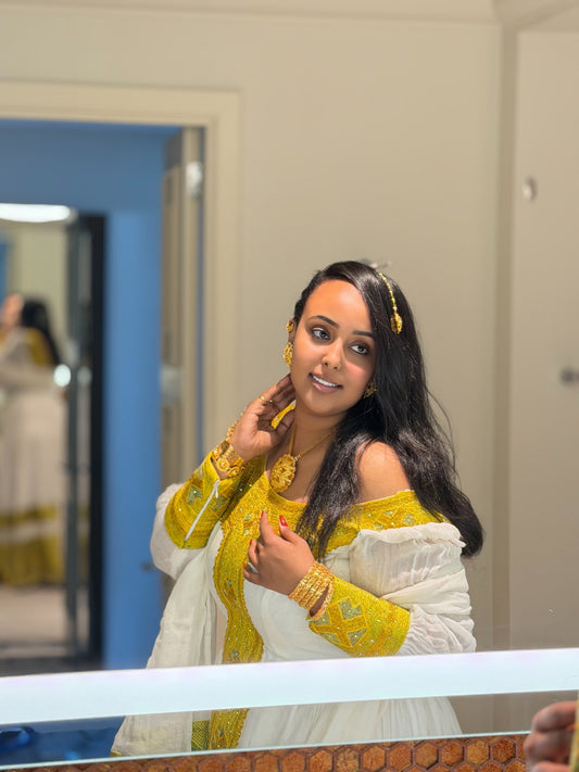 A woman wearing a traditional off-the-shoulder yellow and white Ethiopia Clothes with gold jewelry, posing in front of a mirror in an indoor setting.