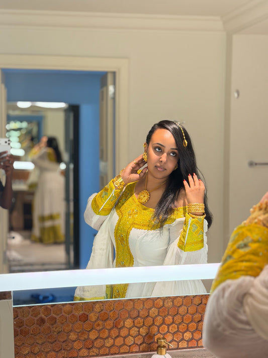 A woman wearing a traditional white and gold embroidered Ethiopia Clothes stands in front of a bathroom mirror, adjusting her hair and jewelry. The scene is set in a real indoor environment with a tiled countertop and doorway in the background.