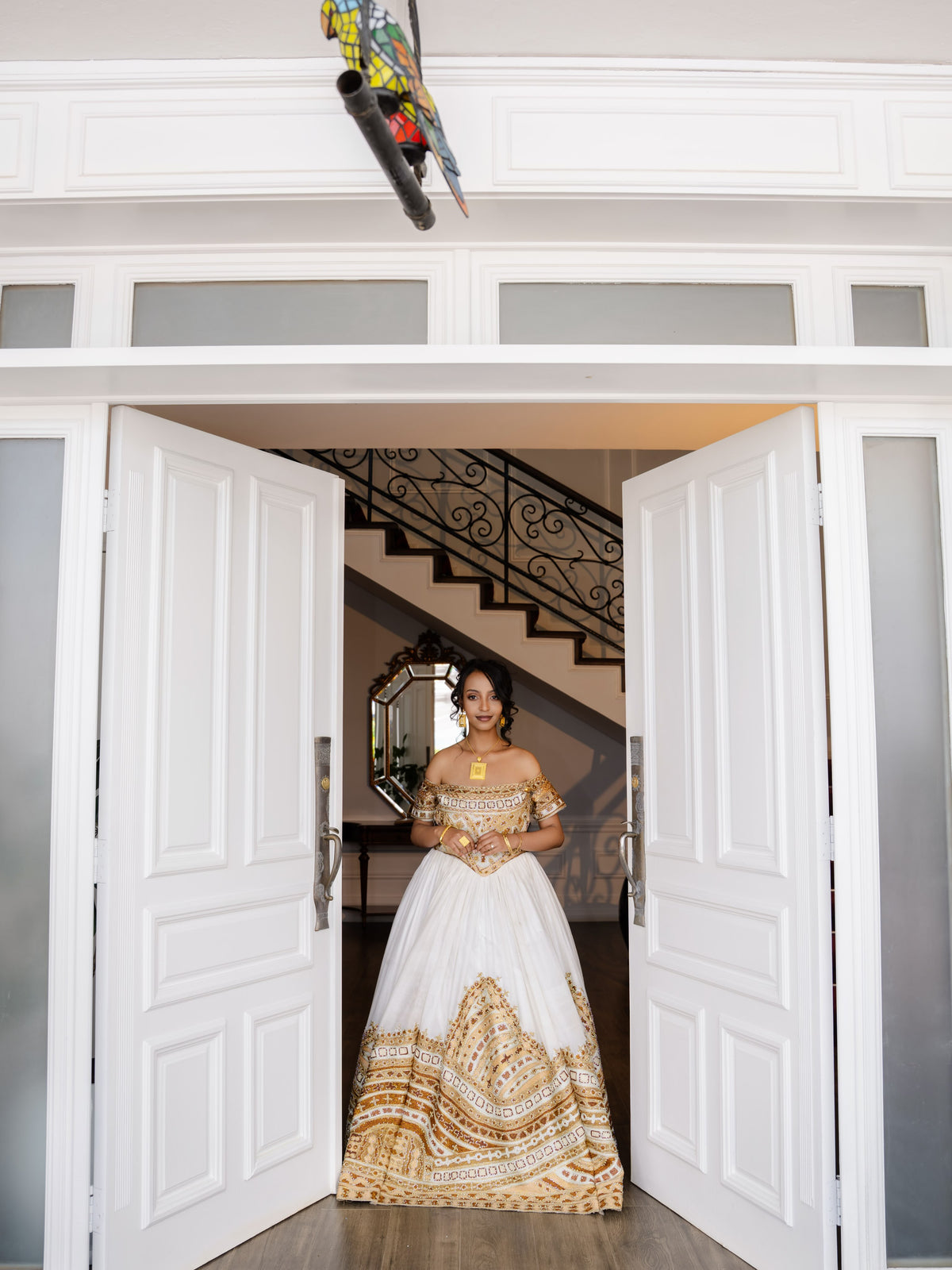 A woman wearing a white ball Habesha bridal Kemis with gold embroidery stands in a doorway, looking directly at the camera. The dress features a structured white bodice and a voluminous skirt with intricate gold patterns.