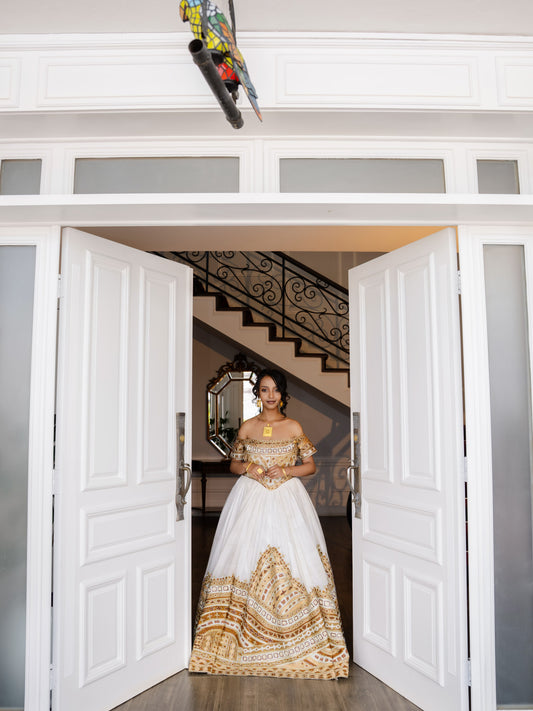 A woman wearing a white ball Habesha bridal Kemis with gold embroidery stands in a doorway, looking directly at the camera. The dress features a structured white bodice and a voluminous skirt with intricate gold patterns.