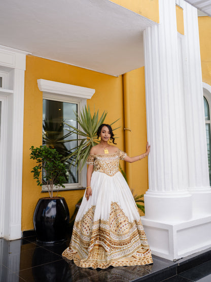 A woman stands on a porch wearing a white off-the-shoulder Habesha bridal Kemis with ornate gold embroidery concentrated on the bodice and lower skirt. The setting features a bright yellow wall, white columns, a black planter with a palm plant, and dark tiled flooring.
