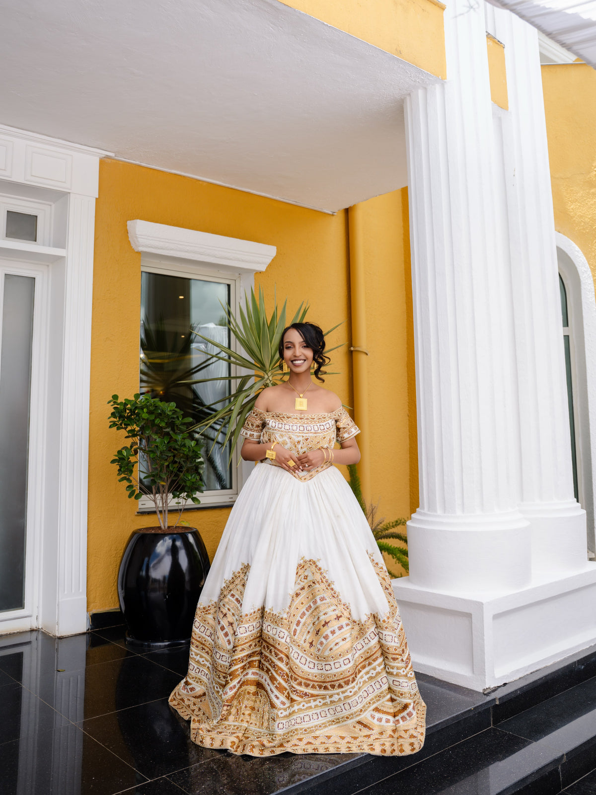 A woman wearing a white ball Habesha bridal Kemis with gold and beige bohemian-style patterns, standing on a black tiled porch in front of a yellow and white building.