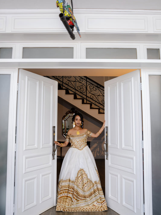 A woman stands in a grand white doorway wearing an off-the-shoulder white Habesha bridal Kemis with extensive gold embroidery concentrated on the bodice and a large gold patterned hem. She is holding open the doors, with a staircase visible in the background.