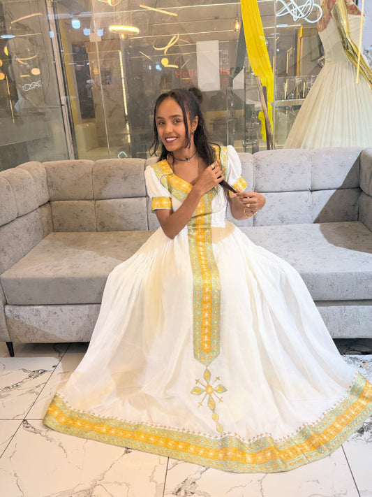 A young woman wearing a white traditional Ethiopia clothes with yellow and green embroidery, sitting on a gray sofa in a boutique setting.