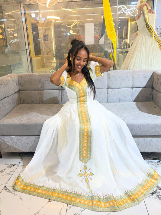 A woman wearing a white Ethiopia clothes with yellow and green embroidery, posing in front of a gray sofa inside a boutique.