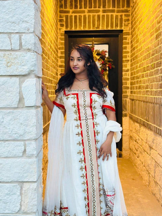 Woman in a white Habesha Kemis with red and gold accents standing in a doorway.