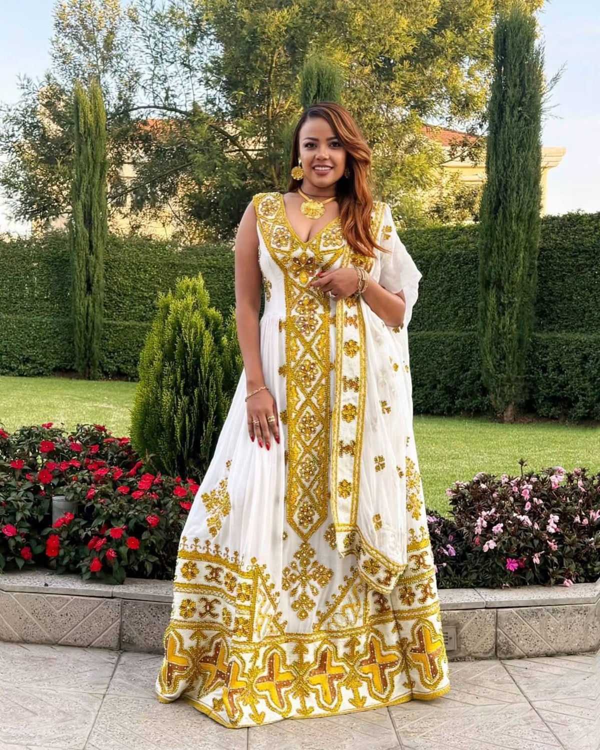 A woman stands outdoors in a garden wearing a white traditional Ethiopian Store with extensive gold embroidery and a matching dupatta.