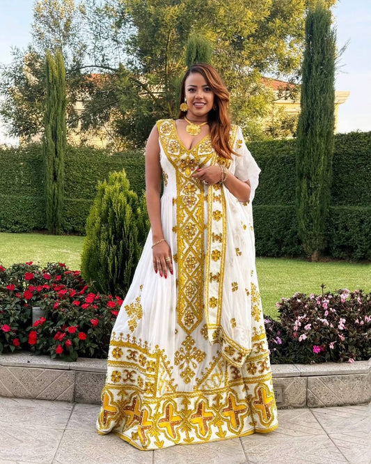 A woman stands outdoors in a garden wearing a white traditional Ethiopian Store with extensive gold embroidery and a matching dupatta.