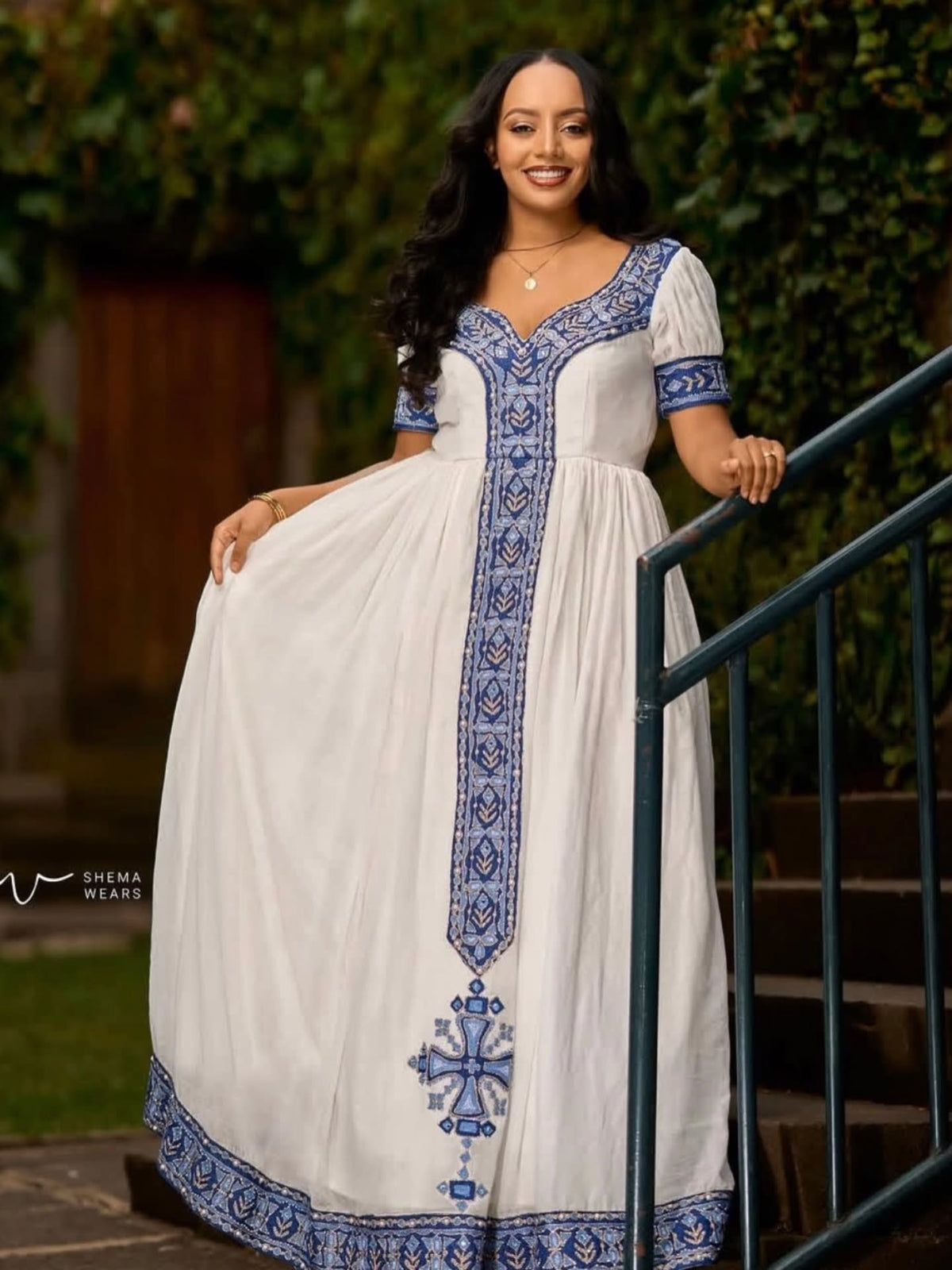 A woman wearing a white maxi Habesha Kemis with blue embroidery, standing on outdoor steps next to a green railing.