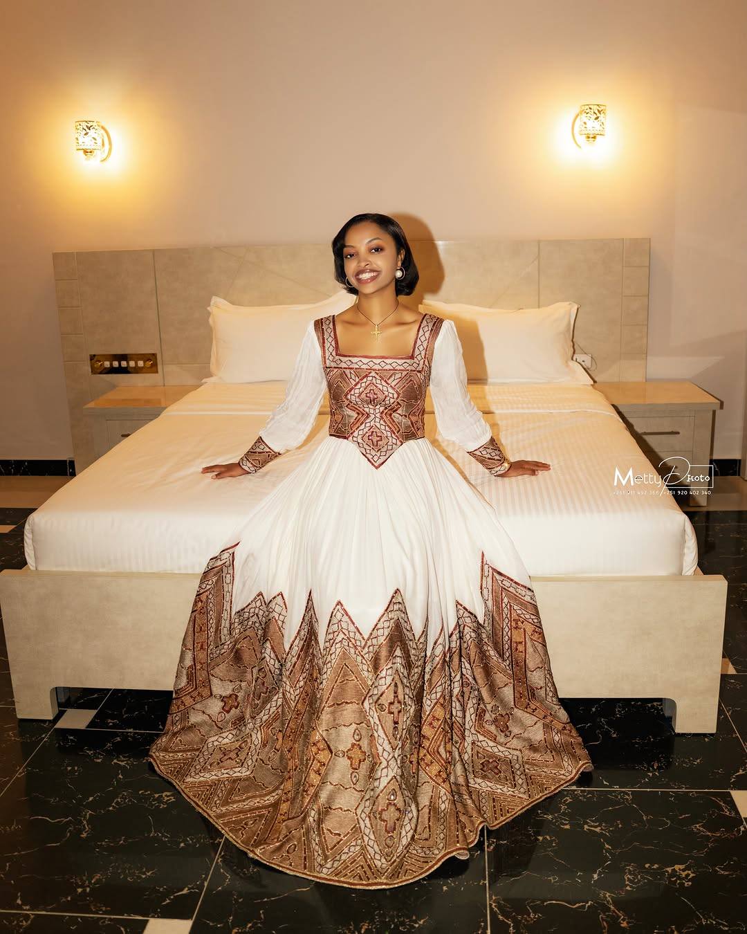 A woman wearing a traditional Ethiopian Traditional Clothingwith a white skirt and brown patterned bodice and hem, sitting on a bed in a hotel room.