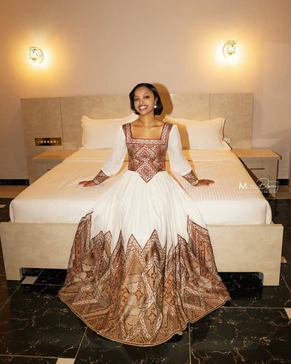 A woman wearing a traditional Ethiopian Traditional Clothingwith a white skirt and brown patterned bodice and hem, sitting on a bed in a hotel room.