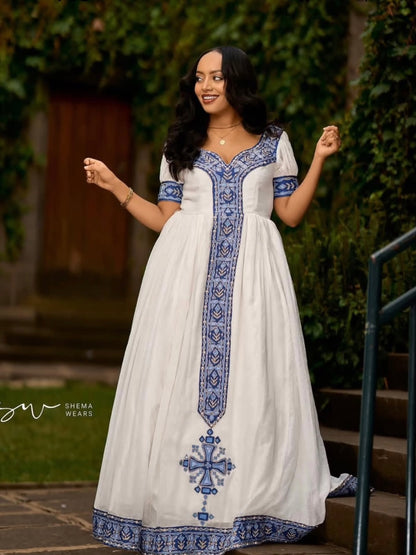 A woman wearing a white maxi Habesha Kemis with blue embroidery, standing on outdoor steps next to a green railing.