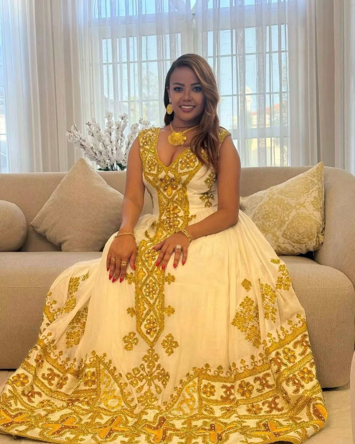 A woman wearing a cream and gold heavily embroidered traditional Ethiopian Store sits on a beige sofa in a bright living room setting.