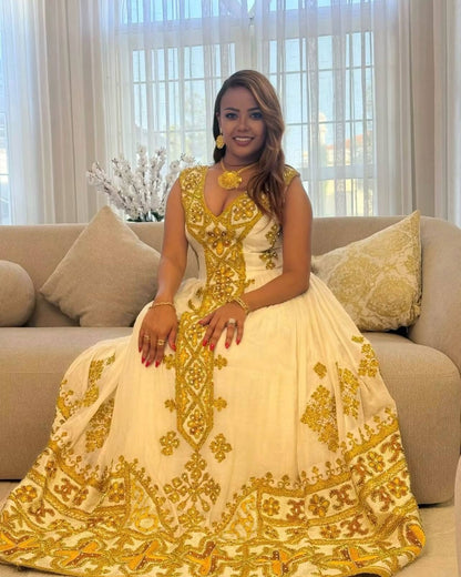 A woman wearing a cream and gold heavily embroidered traditional Ethiopian Store sits on a beige sofa in a bright living room setting.