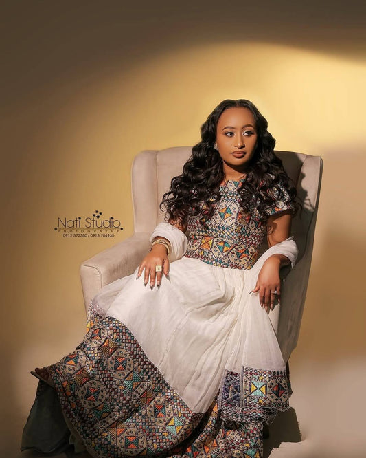 A woman seated in a beige armchair wearing a long white Habesha Dress  with a colorful geometric patterned bodice and hem, photographed against a warm beige studio background. A 'Nait Studio' watermark is visible on the left.