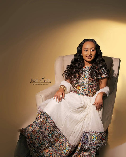 A woman with long, wavy black hair sits in a beige Habesha Dress , wearing a white traditional dress with colorful geometric patterns on the bodice and hem. The photo is taken in a studio with a warm, spotlighted beige background, and a watermark for 'Nail Studio' is visible on the left.