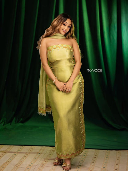 A woman modeling a strapless olive green traditional Habesha dress  with gold embroidery and a matching draped scarf, standing against a dark green curtain backdrop.