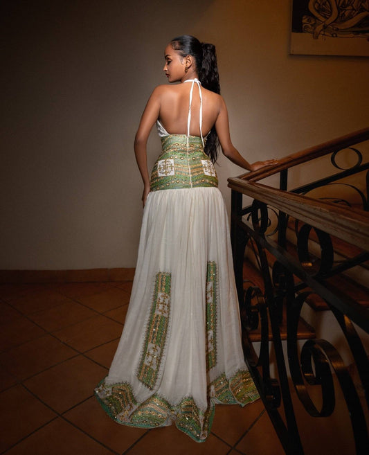 A woman wearing a white halter maxi Ethiopian traditional clothing with green and gold embroidered details, shown from behind as she stands on a tiled staircase landing next to a wooden railing.