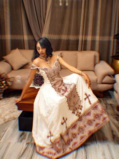 A woman wearing a white off-the-shoulder Ethiopia Clothes with brown and beige embroidered patterns, sitting on a wooden bench in a living room setting.