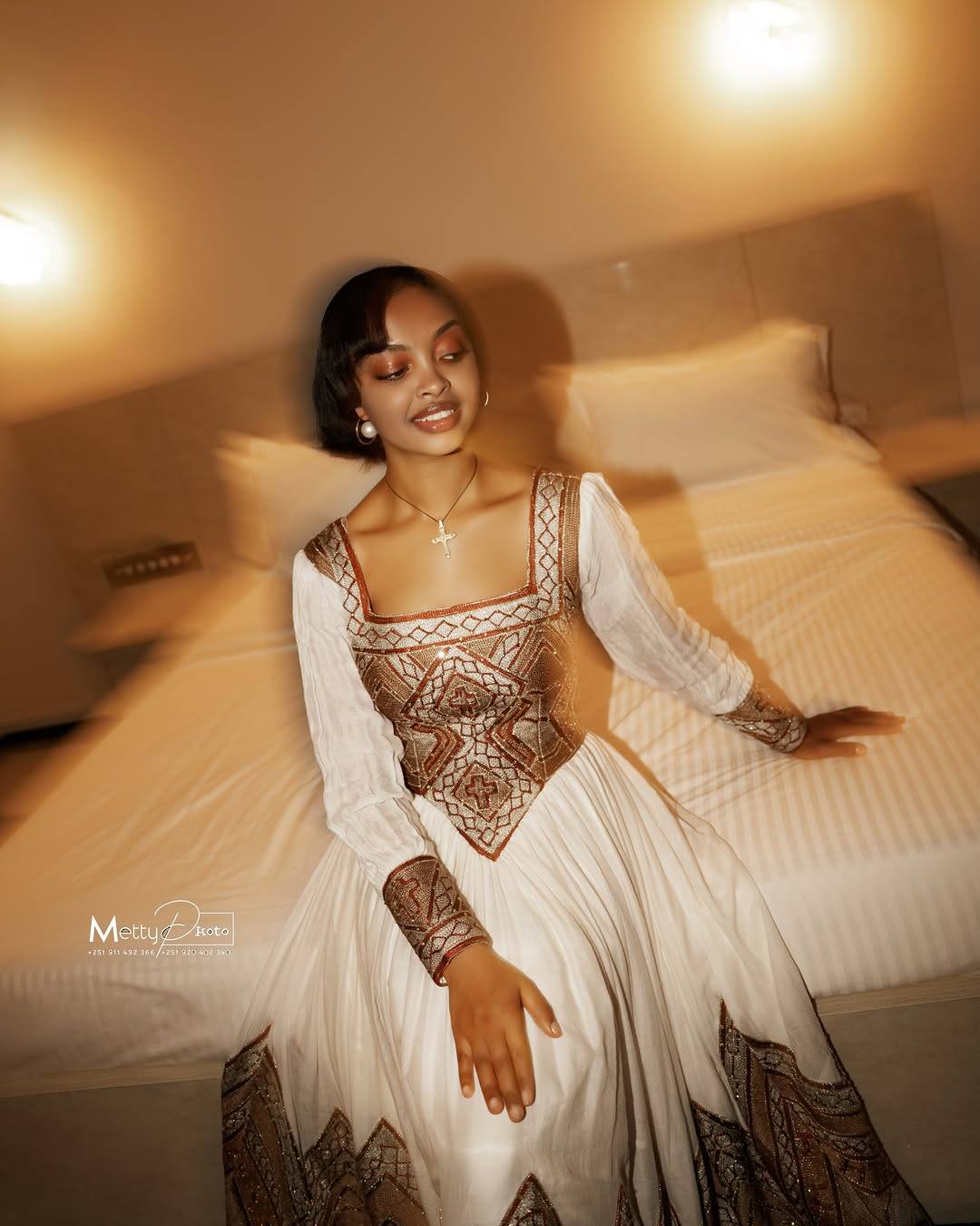 A woman wearing a long white Ethiopian Traditional Clothing with a bronze lace corset bodice and a bronze lace flared skirt sits on a bed in a warmly lit bedroom.