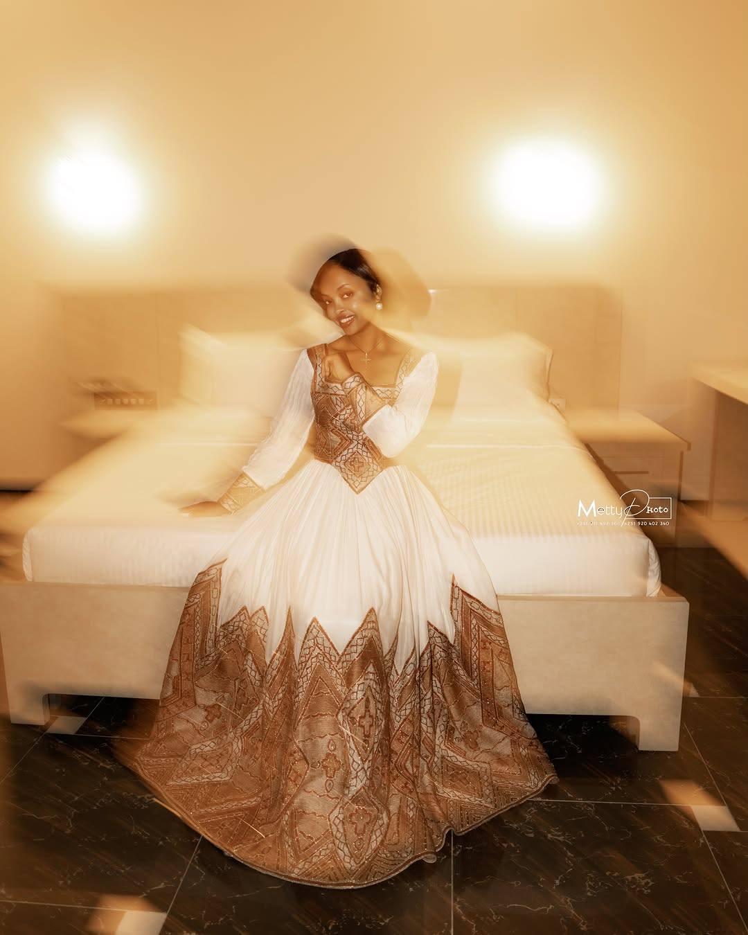 A woman wearing a long white Ethiopian Traditional Clothing with a bronze lace corset bodice and a bronze lace flared skirt sits on a bed in a warmly lit bedroom.