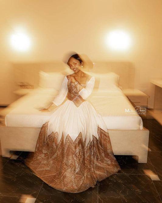 A woman wearing a long white Ethiopian Traditional Clothing with a bronze lace corset bodice and a bronze lace flared skirt sits on a bed in a warmly lit bedroom.