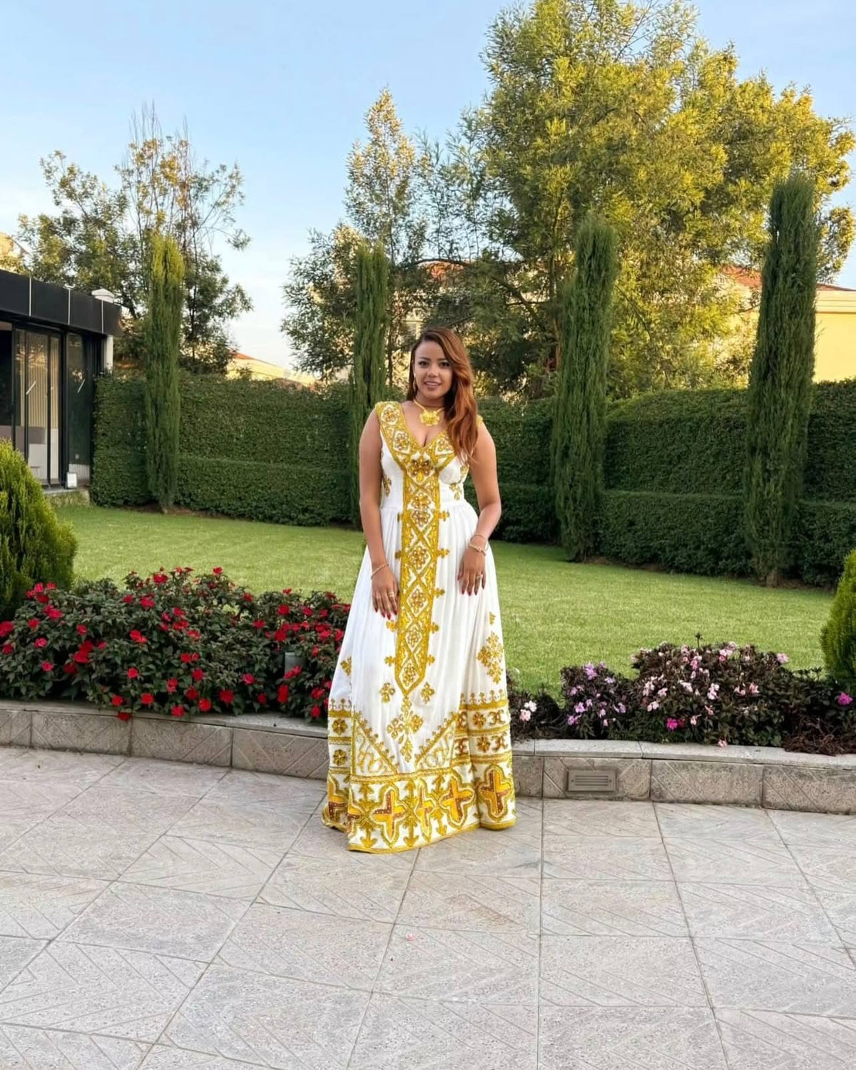 A woman wearing a sleeveless white maxi Ethiopian Store with gold embroidery stands on a stone patio in front of a landscaped garden with green hedges, trees, and flower beds.