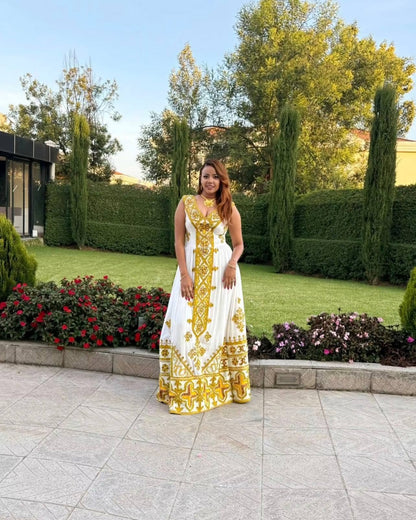 A woman wearing a sleeveless white maxi Ethiopian Store with gold embroidery stands on a stone patio in front of a landscaped garden with green hedges, trees, and flower beds.