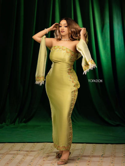 A woman models a floor-length olive green Habesha dress with gold embroidery and matching draped sleeves against a dark green curtain backdrop. The brand name 'TOPAZION' is visible on the right side of the image.