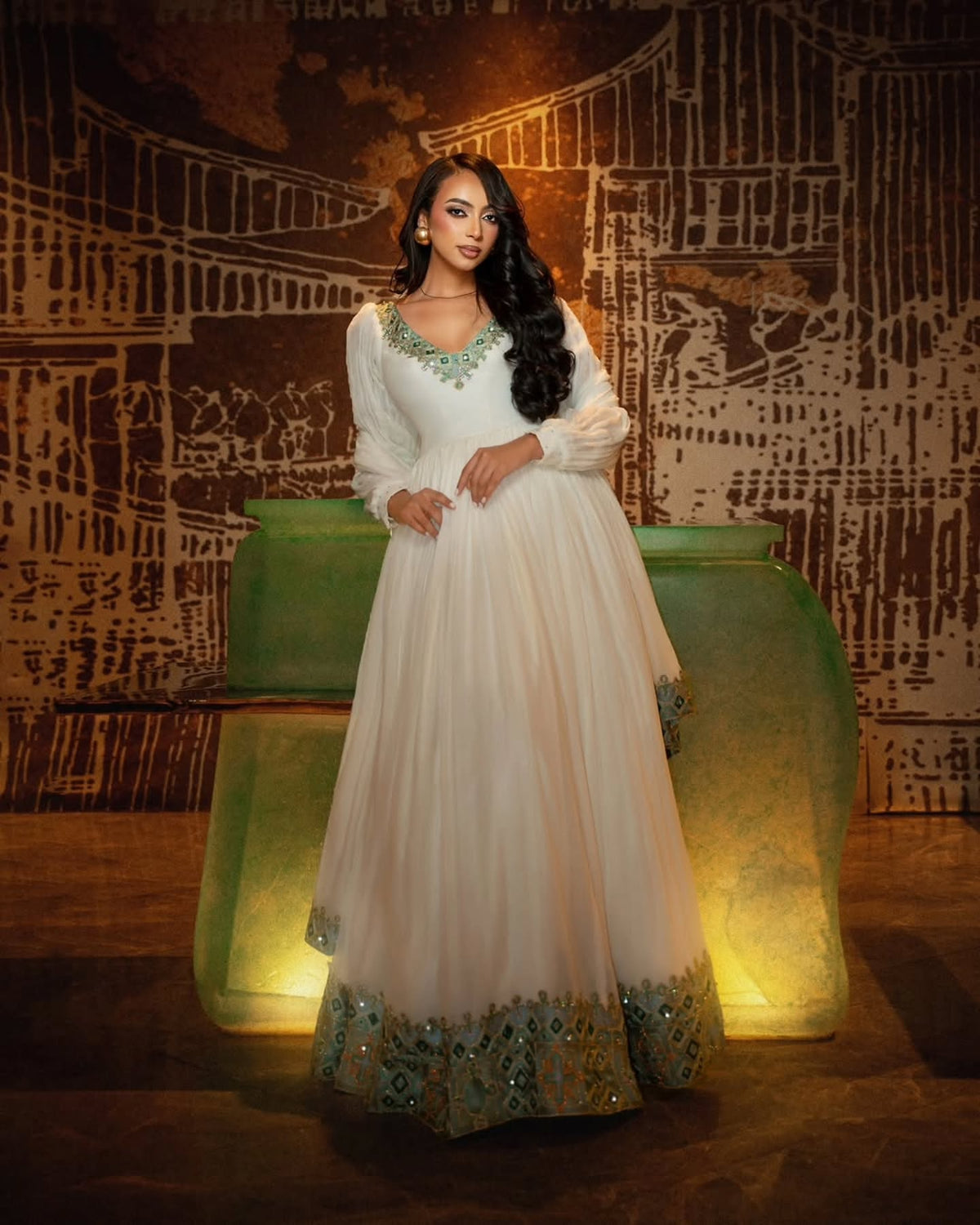 A woman models a long white Anarkali Habesha Dress  with green and gold embroidery on the neckline and hem, standing in front of a stylized green and brown backdrop.