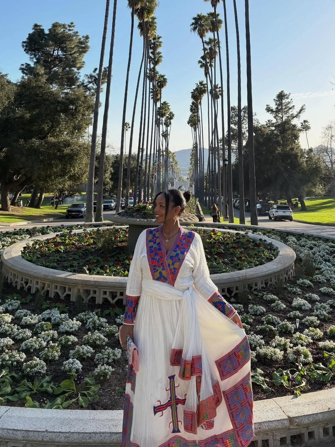 Person wearing a traditional Ethiopian Traditional Dress  with colorful patterns in a park with palm trees and flowers.