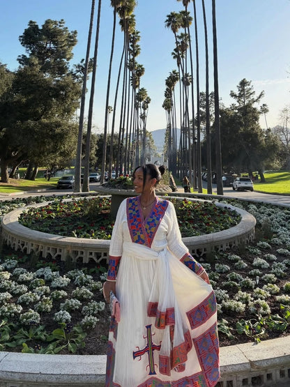 Person wearing a traditional Ethiopian Traditional Dress  with colorful patterns in a park with palm trees and flowers.