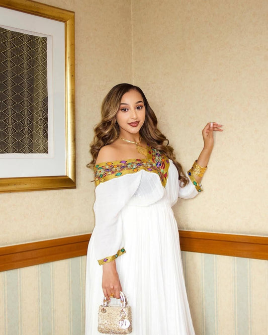 A woman wearing a white off-the-shoulder long-sleeve habesha kemis with colorful embroidered trim on the neckline and cuffs, holding a small metallic handbag, standing indoors against a beige patterned wall.