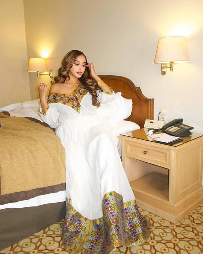 A woman wearing a white Habesha Kemis with colorful floral embroidery sits on a bed in a hotel room. The dress features a white body with ornate multicolor patterns along the neckline and hem.