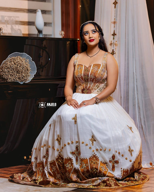 Woman sitting in a white and gold traditional Habesha wedding Kemis featuring a corset bodice and intricate cross embroidery along the hem.