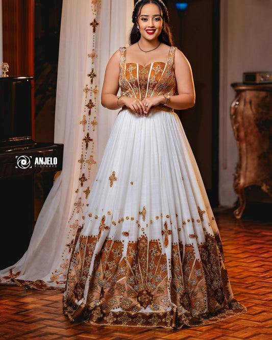 A woman wearing a traditional white and gold embellished Habesha wedding Kemis with a sheer embroidered dupatta, standing in an indoor setting with wooden floors and furniture.