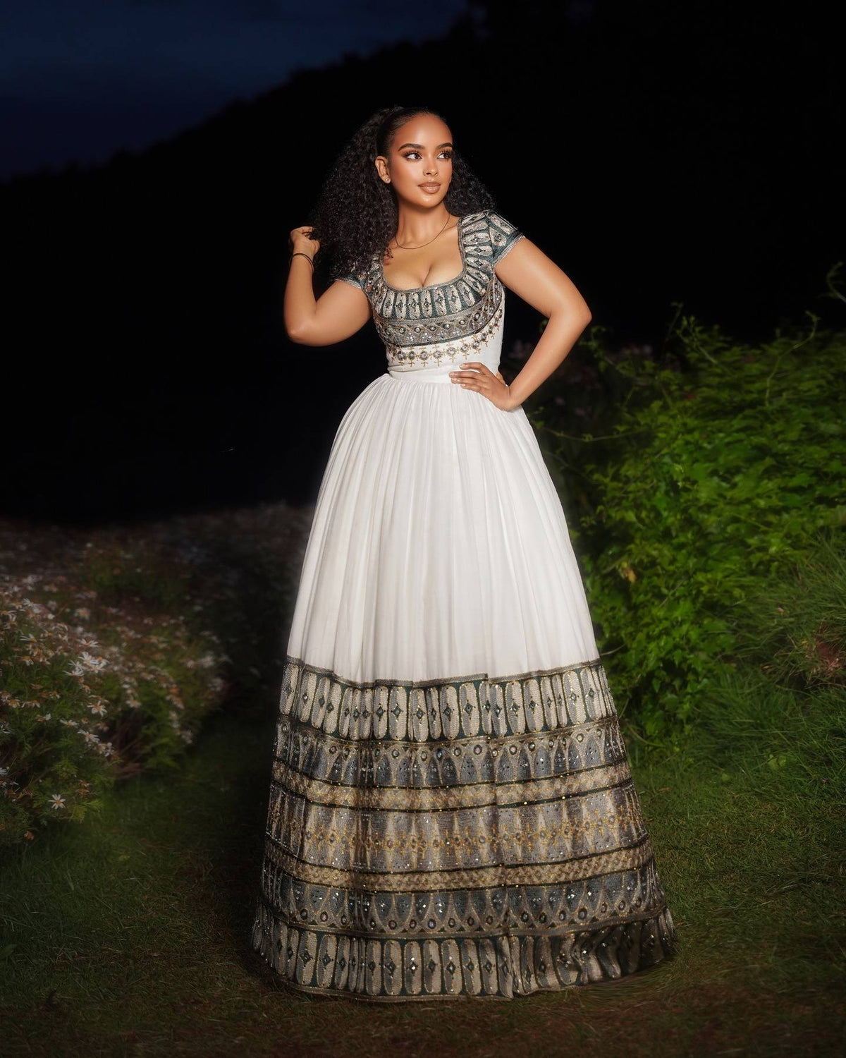 Woman wearing a white and patterned ethiopian dress standing outdoors at night.