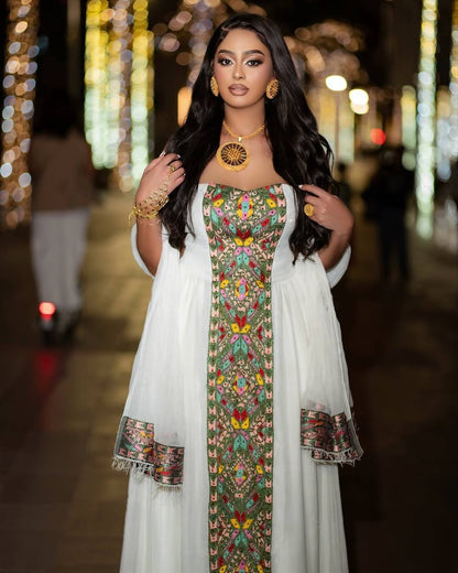 A woman wearing a white traditional habesha kemis with a colorful embroidered floral panel down the front and matching embroidered cuffs, photographed at night on a softly blurred street with lights in the background.