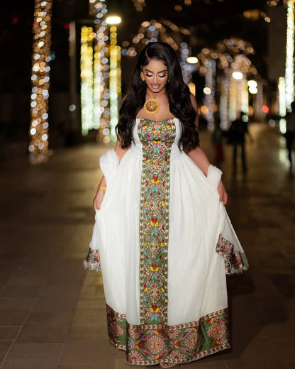 A woman wearing a white traditional habesha kemis  with a colorful embroidered border and a matching white shawl, standing in a nighttime street setting with blurred lights in the background.