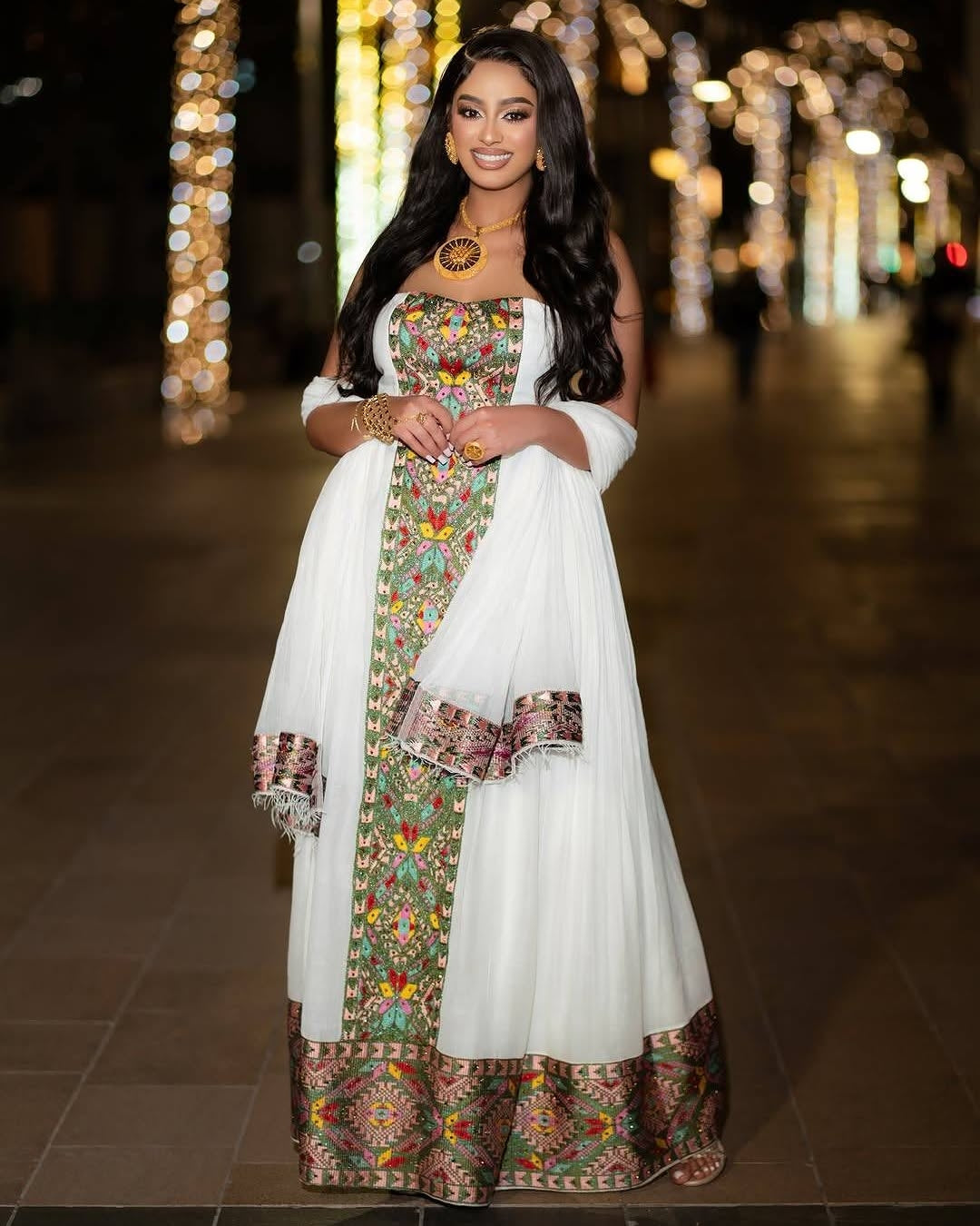 A woman stands outdoors at night wearing a white traditional habesha kemis  with colorful floral embroidery and a matching sheer dupatta draped over her shoulders. The background shows blurred warm lights along a walkway.